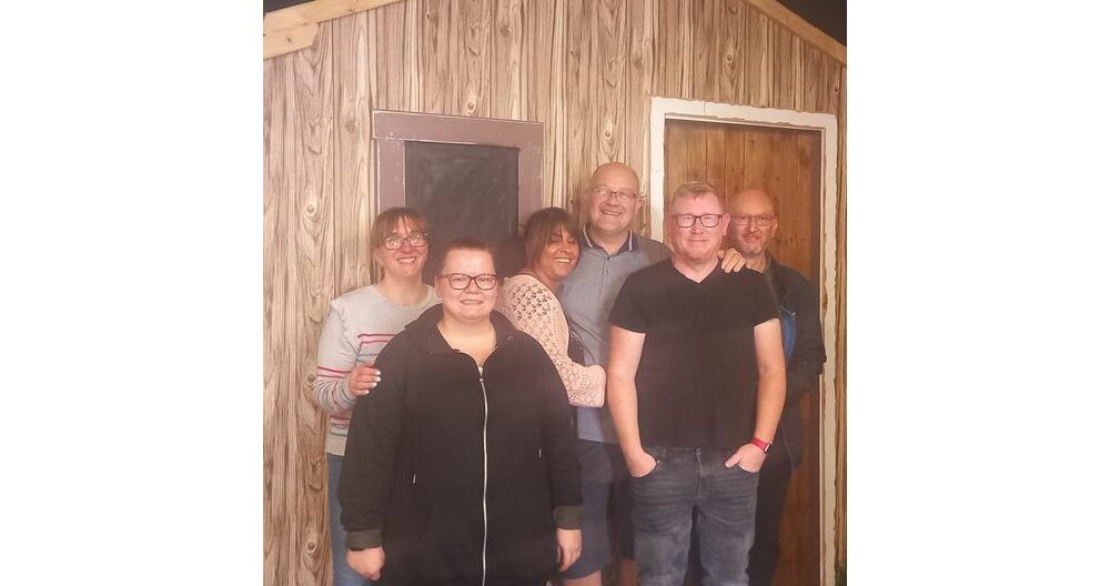 Three men and three women smiling in front of a wooden door and wall.