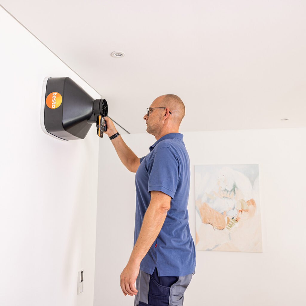 A man in grey trousers and a blue polo t-shirt holds is using the Testo 417 Vane Anemometer with a funnel to measure an outlet high on a wall close to the ceiling.