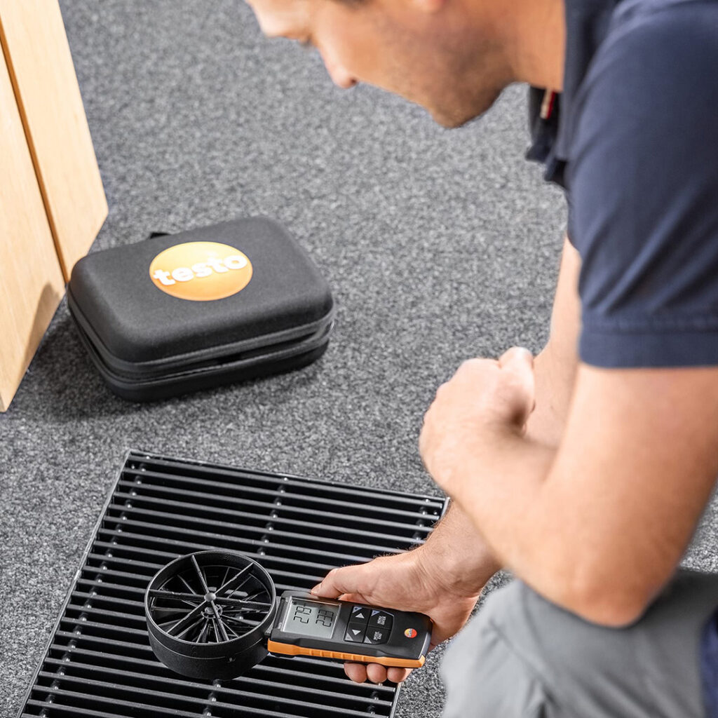 A man in grey trousers and a blue polo t-shirt holds the Testo 417 Vane Anemometer over a ground ventilation grate. 