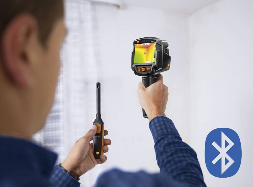 A man in a blue checked shirt is using the Testo 872s Thermal Camera with the Testo 605i Thermohygrometer to assess the mould risk in a room. 