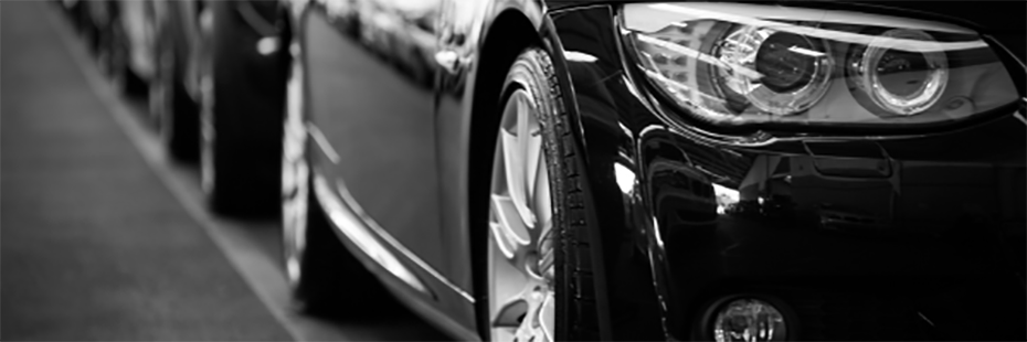 A black and white image of a line of cars. The image focuses on the headlights and sides of the vehicles. 