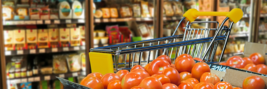 A grocery store. In the forefront are boxes of tomatoes. Behind the tomatoes is a shopping trolly and in the background is a wall of refrigerators containing various produce.   