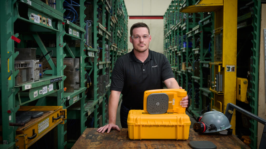 A man in a black polo t-shirt and safety glasses is standing in a warehouse with high green metal shelving either side. On a wooden table in front of him is a ii915 Acoustic Camera on top of a yellow hard case. Next to it is a grey hard hat and ear muffs.  