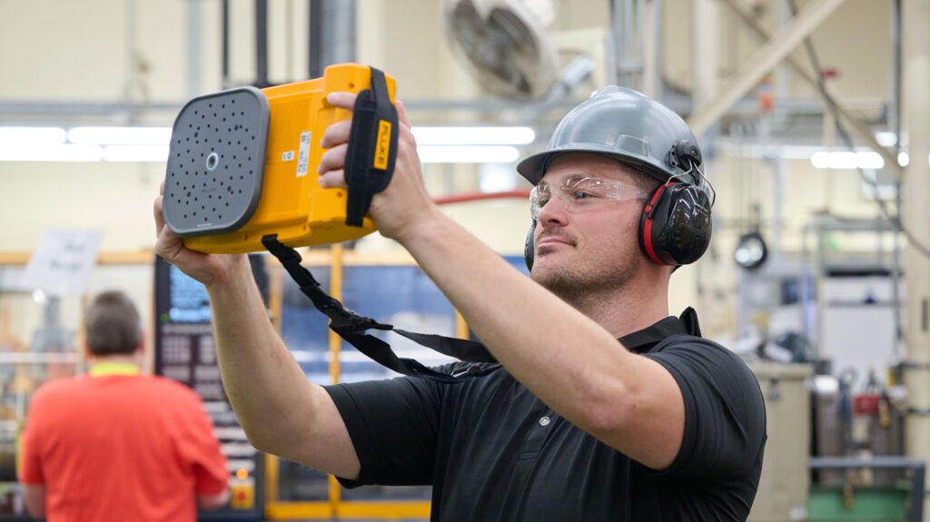 A man in an industrial setting wearing a grey hard hat, safety glasses, and ear muffs is holding up a Fluke ii915 Acoustic Camera. The back of the ii915 is visible.