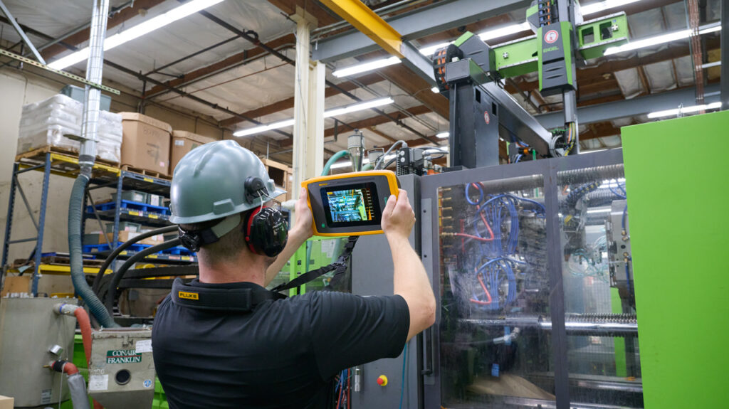 A man in a black t-shirt with a grey hard hat and ear muffs and the ii915 on a neck strap is using the acoustic camera to inspect machinery in a warehouse. The display is visible. A sound map photo is visible on the display. It is showing a fault. 