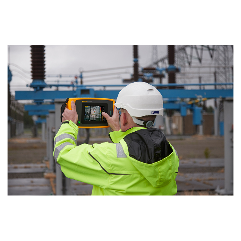 A man in a green high-vis jacket and white hard hat is using a ii915 Acoustic Camera to inspect an utility plant. The display is visible. On the display is a sound map photo showing a PD fault. 