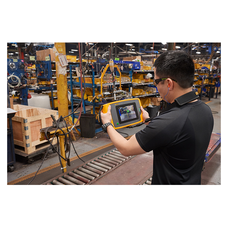 A man in a black t-shirt, safety glasses, and red ear plugs is holding up an ii915 Acoustic Camera which he is wearing around his neck via a neck strap. He is looking at conveyor  in a warehouse. The display is visible. A sound map photo is visible on the display. 