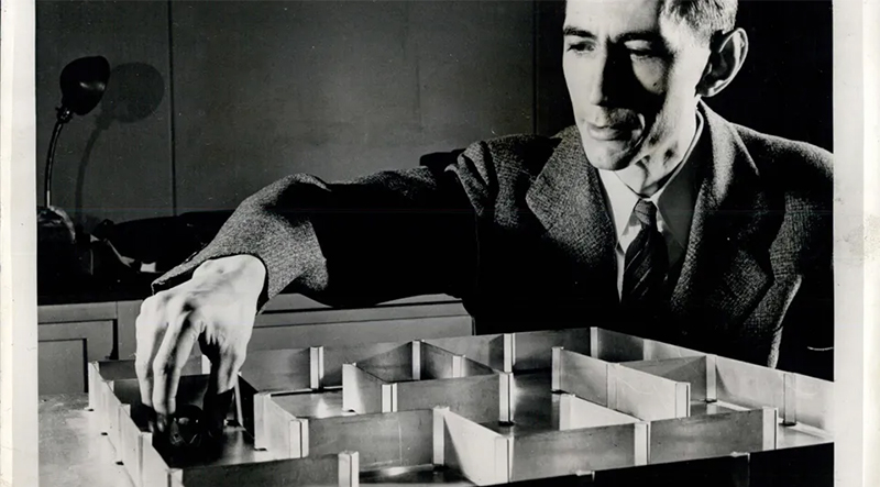 Black and white photo of Claude Shannon, wearing a suit and tie, placing Theseus, an electromechanical mouse, in a small maze. 